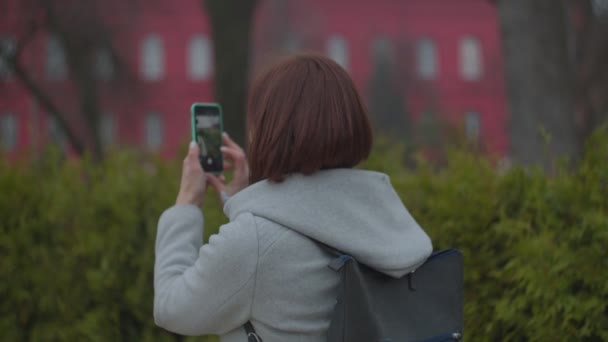 Brunette kvinde tager billede af byens syn på sin smartphone i efteråret park, bakvisning. Kvinde turist i grå frakke med rygsæk gør foto af universitetet . – Stock-video