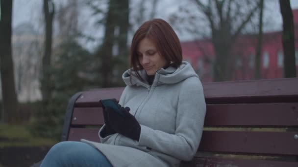 스마트폰을 들고 가을 공원에 벤치에 앉아 있는 갈색 머리 여자 어른. 온라인 서핑을 하며 웃고 있는 여성. — 비디오