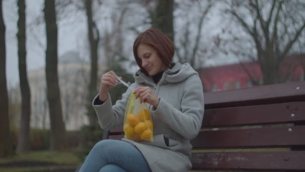 Young brunette woman peeling tangerines on bench in fall park. Female with eco-friendly bag with tangerines smiling while eating mandarines. — Stock Video