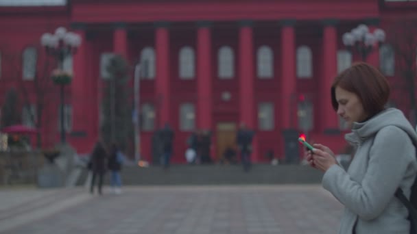Mladá žena se dívá na svůj smartphone s červenou budovou za zády. Žena turista v šedém kabátě s batohem chůze v blízkosti města zraku univerzity. — Stock video