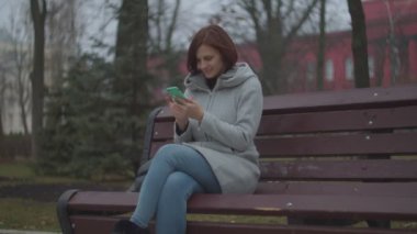 Sonbahar parkında bankta oturan akıllı telefonlu esmer kadın. Telefonda sörf yaparken gülümseyen bir kadın.. 