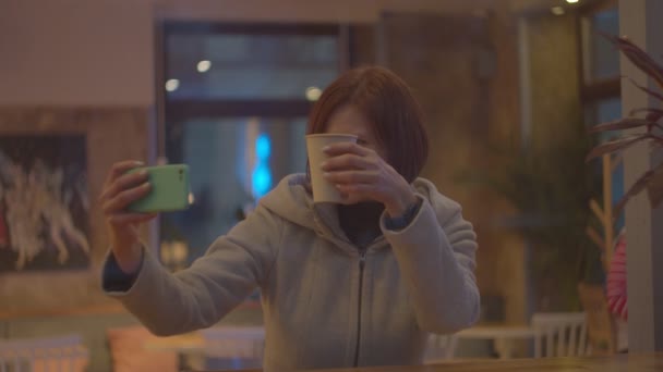 Junge Bloggerin dreht Video mit Smartphone beim Kaffeetrinken im Café, Fensterbild in Zeitlupe. — Stockvideo