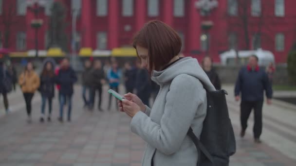 Ung kvinna tittar på sin smartphone med röd byggnad bakom. Kvinnlig turist i grå rock med ryggsäck promenader nära staden syn universitet. — Stockvideo