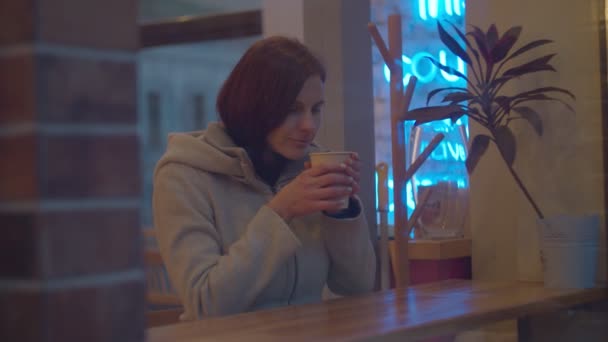 Młode kobiety w płaszczu pijące kawę w kawiarni, strzelające w zwolnionym tempie. Kobieta siedząca przy oknie w kawiarni z filiżanką napoju. — Wideo stockowe