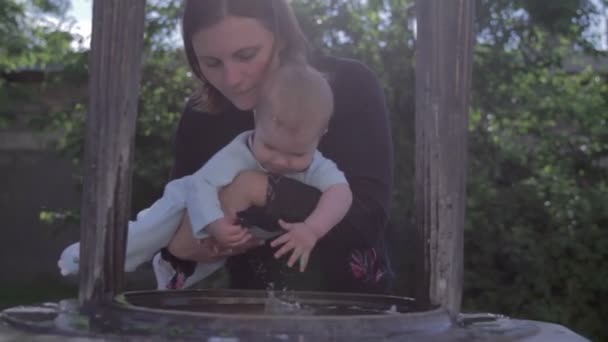 Young mother holding newborn baby boy over the well. Kid in moms hands touching water with drops. — Stock Video