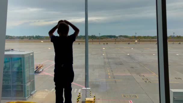 Big boy standing by the airport window, small boy running to join him. Kids watching airplane runway. Children silhouette in airport. — Stock Video