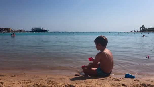 Peuter brunette jongen zitten op de kust en spelen met zand en speelgoed. Broer in opblaasbare ring rennend naar zee. — Stockvideo