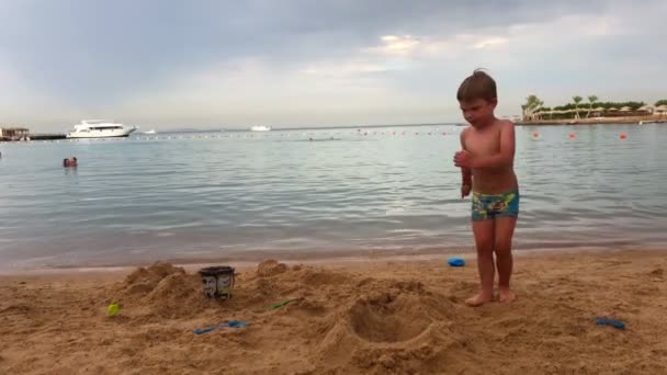 Kleinkind in Badehose läuft am Ufer des Meeres und sitzt im Sand. Kind spielt auf der Meerseite mit Wasser und kann. — Stockvideo