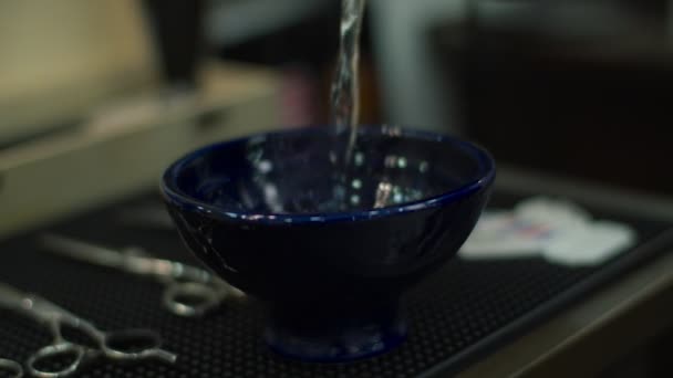 Agua vertiendo en el tazón azul en la estera de goma con el logotipo de la barbería en cámara lenta. Diferentes herramientas de peluquería sobre la mesa . — Vídeos de Stock