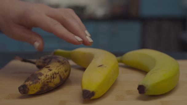Vrouwelijke hand kiezen een van de drie bananen met verschillende rijpheid podium op houten snijplank in slow motion. Vrouwen die groene bananen selecteren. — Stockvideo