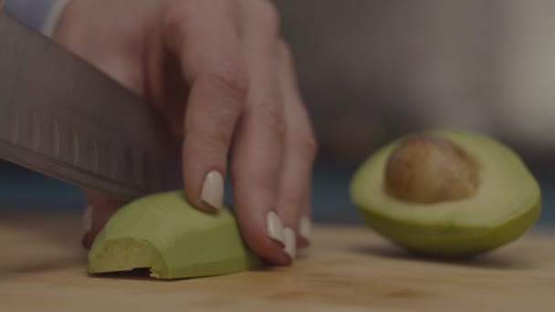 Frauenhände schneiden Avocado mit einem großen Küchenmesser in Zeitlupe auf einem Holzschneidebrett. halbe Avocado mit Kern. — Stockvideo