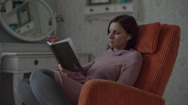 Mujer joven leyendo libro de papel sentado en acogedor sillón naranja en el dormitorio. Lectura femenina y páginas giratorias en libro en cámara lenta . — Vídeo de stock