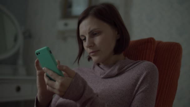 Młoda brunetka dorosłych z bezprzewodowymi słuchawkami surfing online w smartfonie siedzi w przytulnym pomarańczowym fotelu w sypialni. Zamknij się w zwolnionym tempie.. — Wideo stockowe