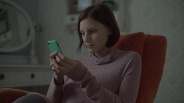 ベッドルームで居心地の良いオレンジのアームチェアに座ってスマートフォンでオンラインサーフィンワイヤレスイヤフォン付きの若い女性のブルネットの大人。スローモーションで閉じます. — ストック動画