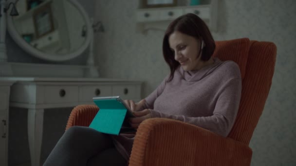 Young smiling female brunette adult with wireless earphones surfing online in tablet computer sitting in cosy orange armchair in bedroom. — Stock Video