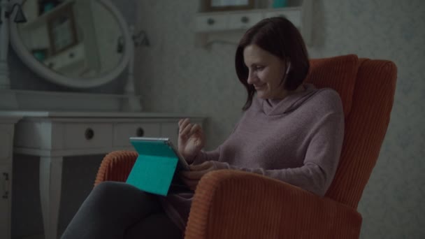 Young female brunette adult with wireless earphones talking online with friend sitting in cosy orange armchair in bedroom. Woman waving hand as greeting to screen. — Stock Video