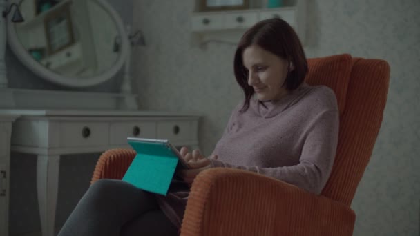 Jonge glimlachende vrouw brunette volwassene met draadloze oortelefoons surfen online in tablet computer zitten in gezellige oranje fauteuil in slaapkamer. — Stockvideo