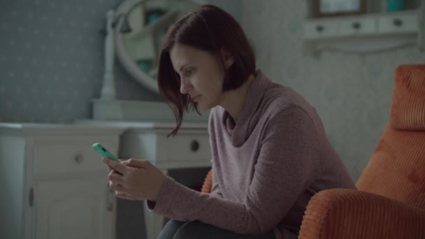 Jonge vrouwelijke brunette volwassene met draadloze oortelefoons surfen online in smartphone zitten in gezellige oranje fauteuil in de slaapkamer. Sluiten in slow motion. — Stockvideo