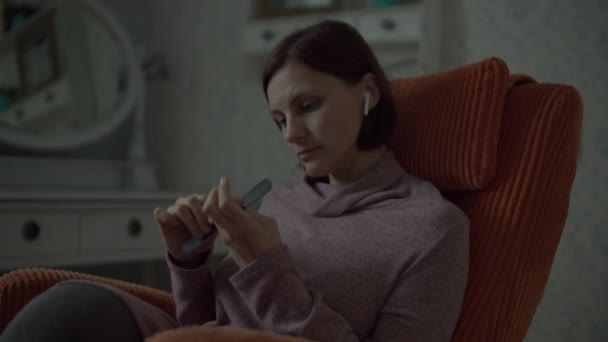 Close up of Young female brunette with wireless earphones using nail file for home manicure sitting in cosy orange armchair in bedroom. — Stock Video