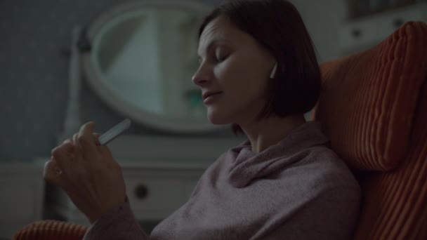 Close up de Jovem morena feminina com fones de ouvido sem fio usando arquivo de unhas para manicure em casa sentado em poltrona laranja acolhedora no quarto . — Vídeo de Stock