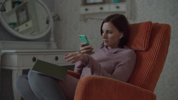 Fiatal nő olvasás papír könyv és fényképezés oldal okostelefon, hogy ellenőrizze a kérelmet. Barna cella és könyv ül a hangulatos narancssárga fotel a hálószobában — Stock videók