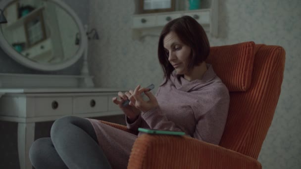 Молодая брюнетка с беспроводными наушниками с помощью пилка для ногтей для домашнего маникюра сидит в уютном оранжевом кресле в спальне . — стоковое видео