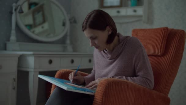 Joven mujer morena pintura para adultos con pincel sentado en acogedor sillón naranja en el dormitorio en cámara lenta . — Vídeos de Stock