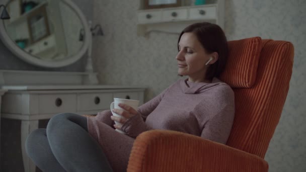 Young brunette woman with wireless earphones and cup of hot drink enjoying the music sitting in cosy orange armchair in bedroom. — Stock Video