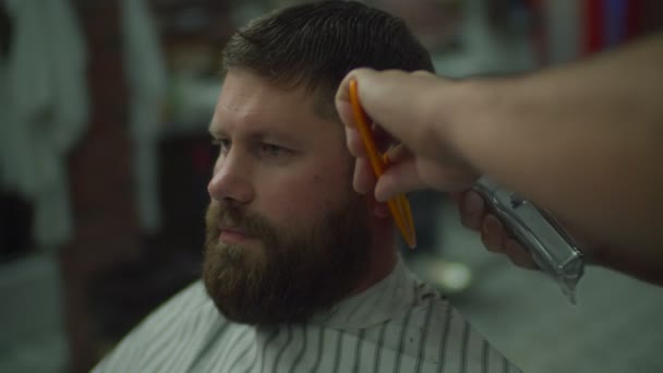 Frisör gör frisyr för skägg man i frisör cape genom elektrisk rakmaskin och kam i frisör butik i slow motion. — Stockvideo