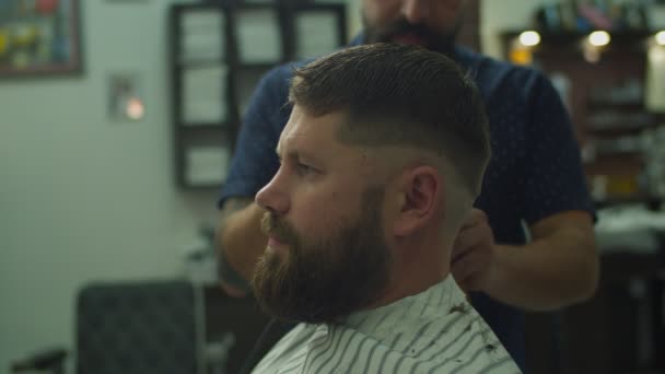 Vista lateral do barbeiro fazendo corte de cabelo para o homem em capa de cabeleireiro na barbearia em câmera lenta . — Vídeo de Stock