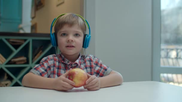 Kleuterschooljongen in kleur koptelefoon eten appel en bewegend hoofd tijdens het luisteren naar de muziek aan tafel. Lachend en lachend kind. — Stockvideo