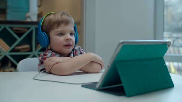 Przedszkolak w kolorowe słuchawki oglądanie komputera tablet przy stole. — Wideo stockowe