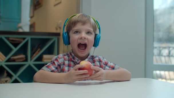 Förskola pojke i färg hörlurar äta äpple och flytta huvudet medan du lyssnar på musik vid bordet. Ungen ler och skrattar. — Stockvideo