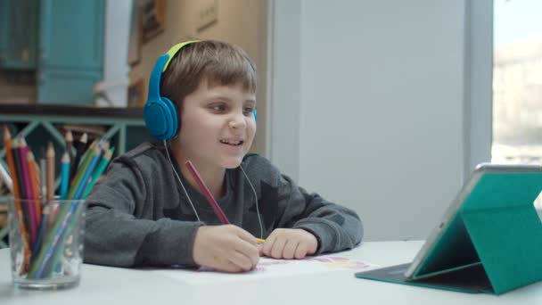Niño de la escuela con autismo en los auriculares a color dibujo con lápices y mirando la pantalla de la tableta. Niño sentado en la mesa y haciendo la tarea . — Vídeo de stock