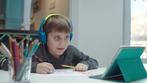 Niño de la escuela con autismo en los auriculares a color dibujo con lápices y mirando la pantalla de la tableta. Niño sentado en la mesa y haciendo la tarea . — Vídeos de Stock