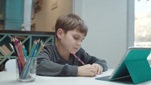 Schüler mit Autismus zeichnen mit Buntstiften und schauen auf den Bildschirm des Tablet-Computers. Kind lächelt und sitzt am Tisch und macht Hausaufgaben. — Stockvideo