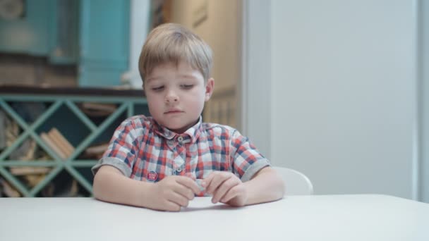 Niño preescolar sintiéndose molesto sentado en la mesa y sosteniendo la tarjeta en las manos. Niño en camisa haciendo suspiro triste . — Vídeo de stock