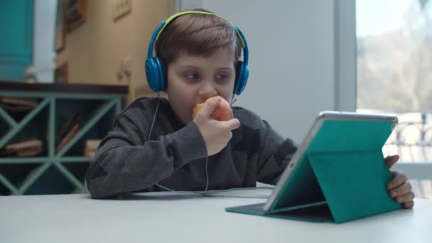 Niño de la escuela con autismo en los auriculares de color comer manzana y jugar en la computadora de la tableta. Niño sentado en la mesa y tocando la pantalla del gadget en cámara lenta . — Vídeos de Stock