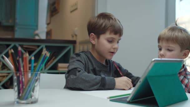 Niño autista dibujando con lápices de color y mirando la tableta en la mesa, su hermano menor viene y se une a la pintura. Hermanos jugando . — Vídeos de Stock