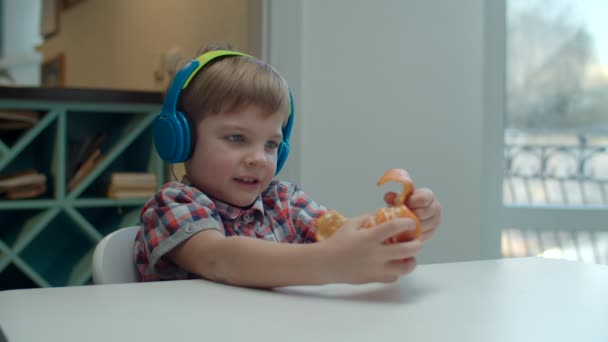 Kleuter in kleur koptelefoon peeling mandarijn en bewegende hoofd tijdens het luisteren naar de muziek aan de tafel. — Stockvideo