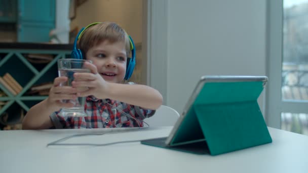 Niño preescolar en auriculares de color viendo tableta y beber vaso de agua en la mesa . — Vídeos de Stock