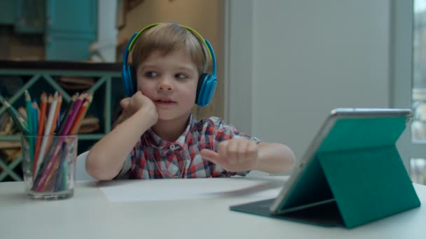 Niño preescolar en auriculares de color mirando el gadget y empezando a dibujar con lápiz de color. Pintura infantil en la mesa . — Vídeos de Stock