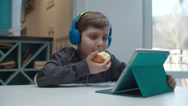 タブレットコンピュータを見ながらアップルを食べ、笑顔でカラーヘッドフォンで自閉症の幸せな学校の少年。子供はテーブルに座ってスローモーションでガジェットを探しています. — ストック動画
