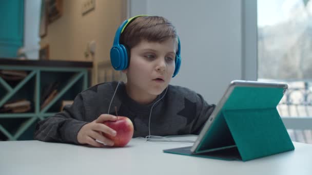 Niño de la escuela con autismo en los auriculares de color comer manzana y jugar en la computadora de la tableta. Niño sentado en la mesa y tocando la pantalla del gadget en cámara lenta . — Vídeos de Stock