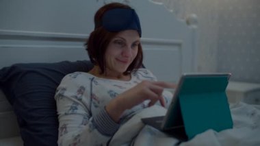 Genç kadın esmer yetişkin yatakta, tablet bilgisayarla internette sörf yapıyor. Kafasında uyku maskesi olan bir kadın alete bakarken gülümsüyor.. 