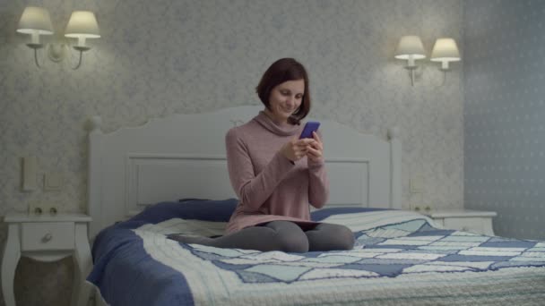 Elbise giymiş esmer kadın internette akıllı telefonuyla yatakta oturuyor. Elinde telefonla gülümseyen bir kadın.. — Stok video