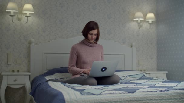 Μπρουνέτ γυναίκα σε φόρεμα surfing σε απευθείας σύνδεση στο φορητό υπολογιστή κάθεται στο κρεβάτι. Γυναίκα χαμογελά στο gadget στα πόδια της. — Αρχείο Βίντεο