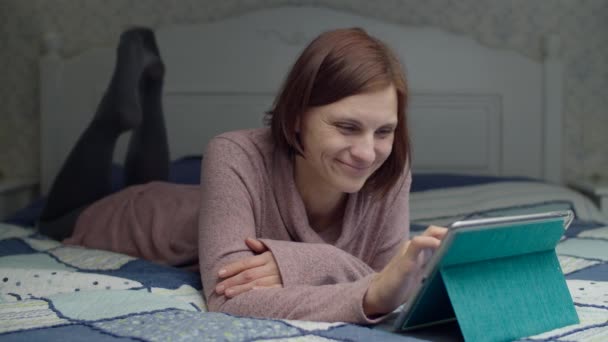 Mujer morena en vestido de surf en línea en la tableta de la computadora acostada en la cama. Mujer balanceando sus piernas y sonriendo en cámara lenta — Vídeo de stock