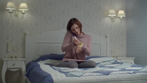 Femme brune en robe parlant par téléphone et hydratant les mains à la crème assise sur le lit. Femme avec téléphone dans les mains souriant — Video