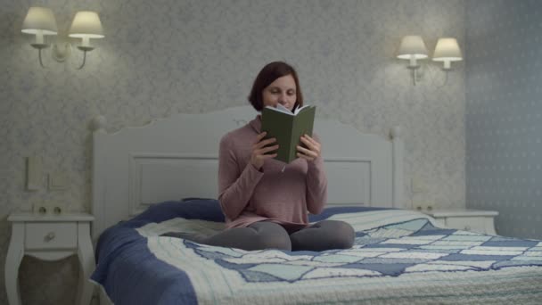 Брюнетка в платье нюхает бумажную книгу, сидя на кровати. Женщина переворачивает страницы в бумажной книге в замедленной съемке . — стоковое видео
